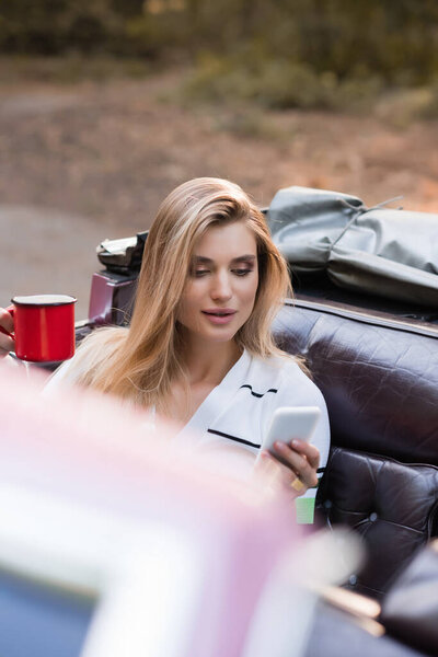 молодая женщина держит чашку кофе и болтает на смартфоне в Cabriolet на размытом переднем плане