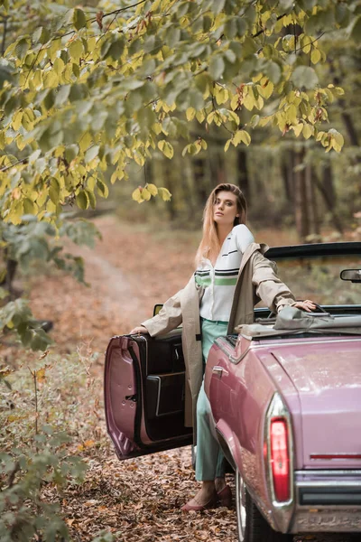 young stylish woman in cape looking at camera while standing near vintage cabriolet in forest