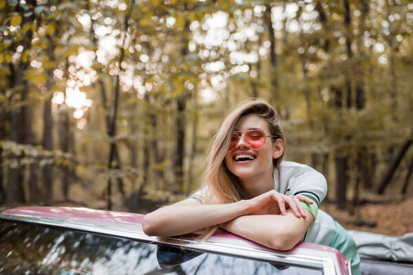 excited woman in sunglasses looking at camera while leaning on windshield of cabriolet