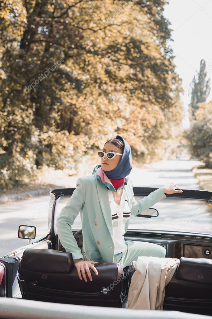 Stylish woman in headscarf looking away on driver seat of roofless car 