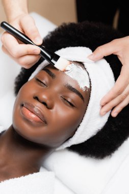 Spa terapisti, spa salonundaki Afrikalı Amerikalı kadının alnına kozmetik fırçasıyla yüz maskesi takıyor.