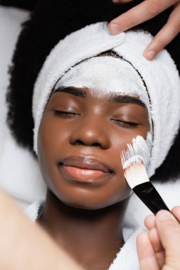 Spa terapisti kaplıca salonunda makyaj fırçasıyla yüz maskesi takarken Afro-Amerikan kadın yalan söylüyor.