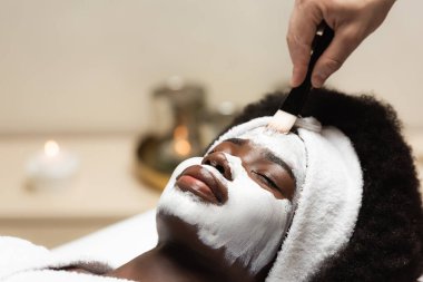 Saç bandı takmış Afrikalı Amerikalı bir kadın spa terapistinin yanında yatıp bulanık arka planda yüz maskesi takıyor. 