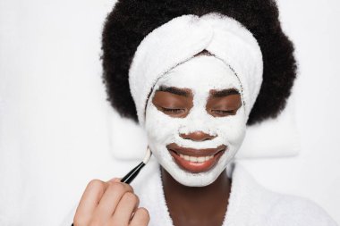Top view of smiling african american woman with face mask near spa therapist with cosmetic brush in spa salon clipart