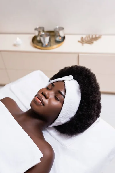 身穿白色头巾 铺有床单的非洲裔美国妇女躺在温泉沙龙的按摩床上 背景模糊不清 — 图库照片