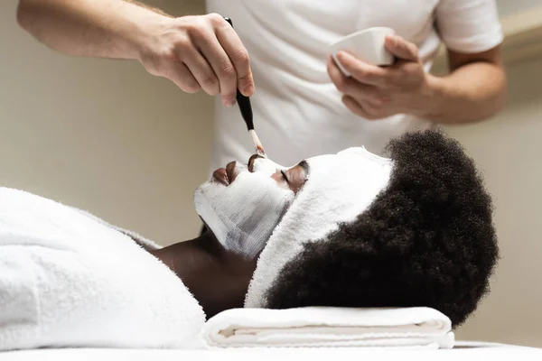 非洲裔美国妇女躺在温泉治疗师身边 在温泉沙龙用化妆品刷鼻子的面罩的侧视图 — 图库照片