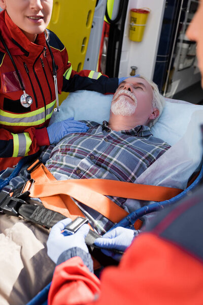 Selective focus of paramedic standing near patient while colleague locking belt of stretcher outdoors 