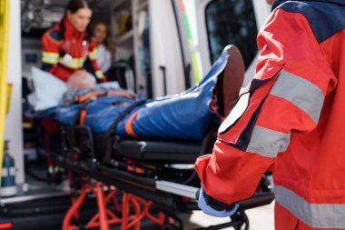 Selective focus of paramedic in uniform carrying stretcher with patient in ambulance ca outdoors  clipart