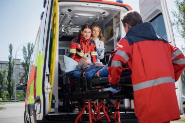 Selective focus of doctor with digital tablet standing in ambulance car near paramedics with patient on stretcher outdoors  clipart
