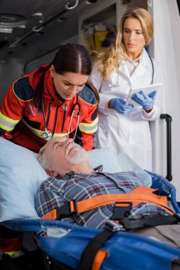 Sağlık görevlisinin yanındaki sedyede yatan yaşlı bir hastanın seçici odak noktası ve ambulansta dijital tabletli bir doktor. 
