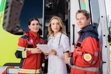 Dijital tabletli doktor sağlık görevlilerinin ve ambulans arabasının yakınındaki kameraya bakıyor. 