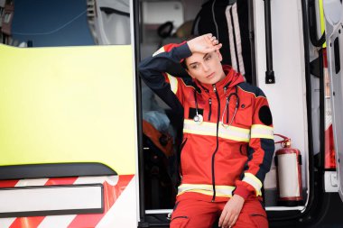 Selective focus of tired paramedic in uniform near ambulance auto outdoors  clipart
