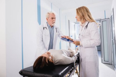 Doktorun not panosuna, kıdemli meslektaşının yanında yazı yazması ve klinikte sedye üzerinde hasta olması seçici bir odak noktası. 