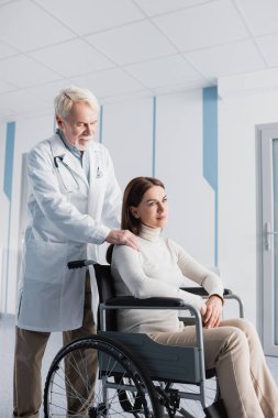 Kıdemli doktor tekerlekli sandalyedeki özürlü hastanın omzuna dokunuyor.