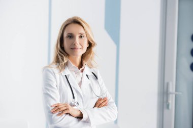 Blonde doctor with stethoscope looking at camera in clinic  clipart