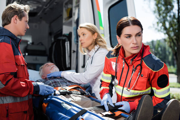 Selective focus of paramedic fasting belt of stretcher near patient, doctor and colleague outdoors 
