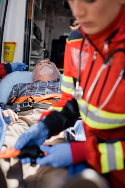 Selective focus of senior man lying on stretcher near paramedics and ambulance car 