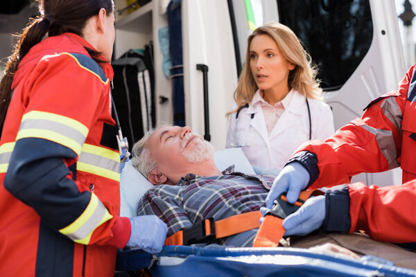 Selective focus of senior man lying on stretcher near paramedics and doctor outdoors 