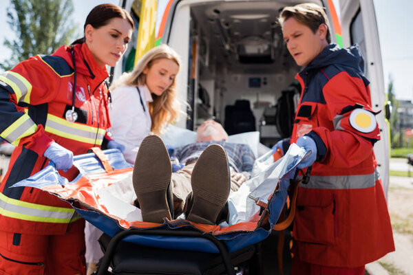Выборочный фокус медиков, держащих носилки рядом с пациентом, врачом и машиной скорой помощи на открытом воздухе 
