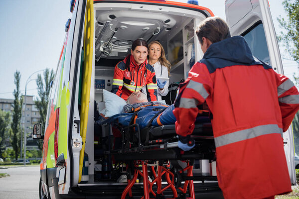 Selective focus of doctor with digital tablet standing in ambulance car near paramedics with patient on stretcher outdoors 