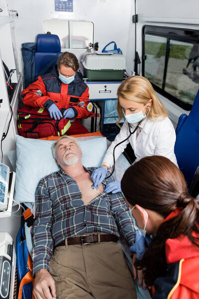 Selective focus of doctor in medical mask examining patient with stethoscope near paramedics in ambulance car 