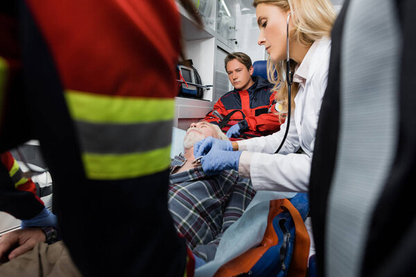 Selective focus of paramedics standing near doctor examining patient with stethoscope in ambulance car 