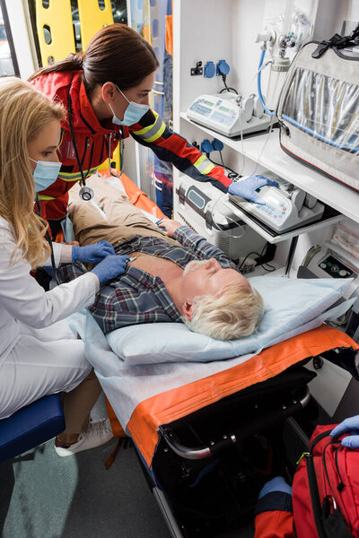 Selective focus of paramedic and doctor in medical masks examining patient with stethoscope in ambulance car 