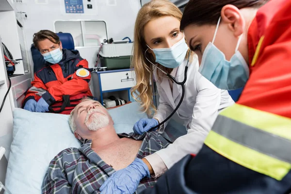 Doktorun steteskoplu seçici odağı tıbbi maskeli sağlık görevlisine ve ambulans arabasındaki hasta hastaya bakıyor. 