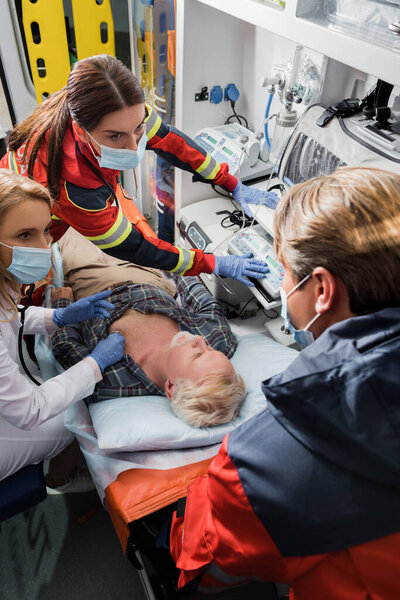 Selective focus of paramedics and doctor in medical masks doing first aid to ill patient in ambulance car 