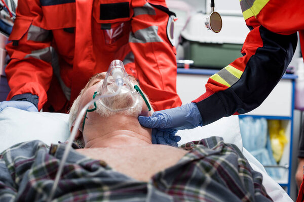 Selective focus of paramedic in latex glove checking pulse of senior patient in oxygen mask near colleague during first aid 