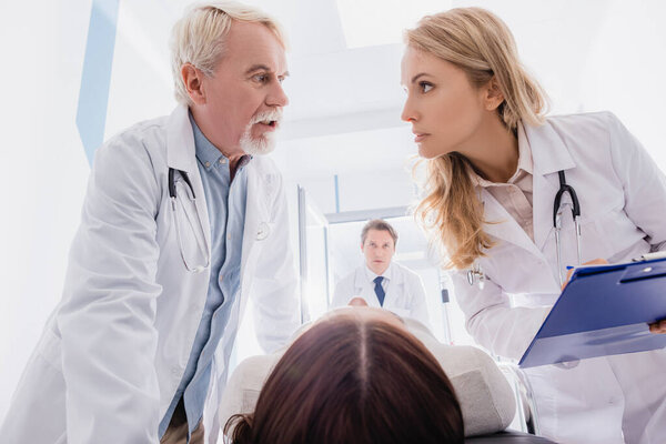 Selective focus of senior doctor talking to colleague with clipboard near patient on stretcher in clinic 