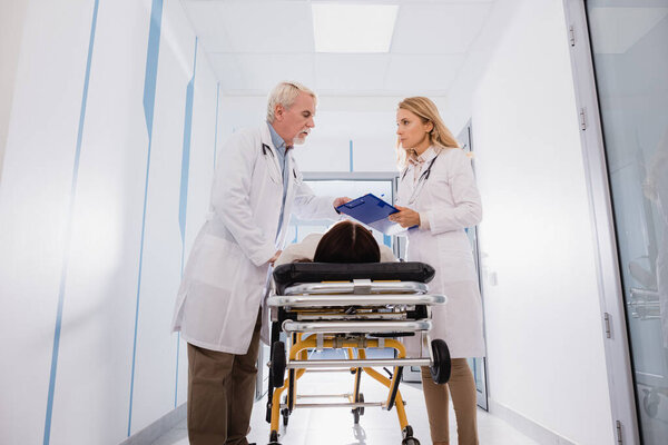 Doctor holding clipboard near senior colleague and sick patient on stretcher in clinic 