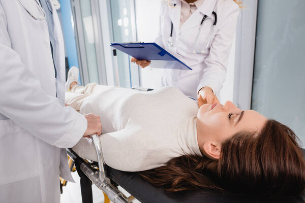 Selective focus of doctor with clipboard checking pulse of patient with closed eyes on stretcher near colleague in clinic 