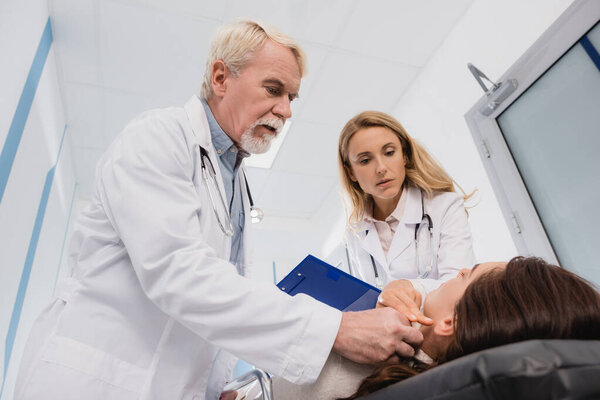 Selective focus of doctors with clipboard checking pulse of woman on stretch in clinic 