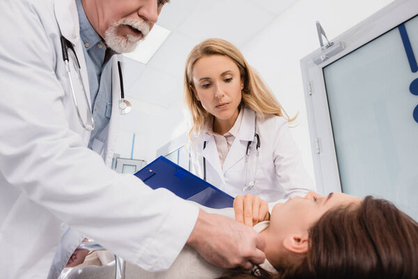 Selective focus of senior doctor checking pulse of patient near colleague with clipboard in clinic 