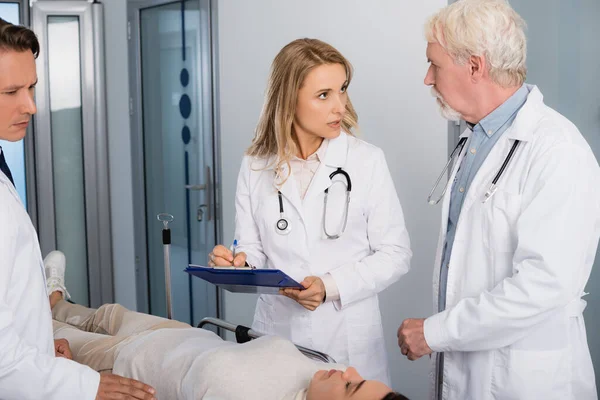 Selective focus of doctor with clipboard talking to senior colleague near patient with closed eyes on stretcher in clinic