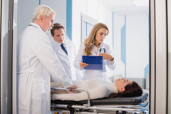 Selective focus of doctors with clipboard looking at patient with closed eyes on stretcher in clinic 