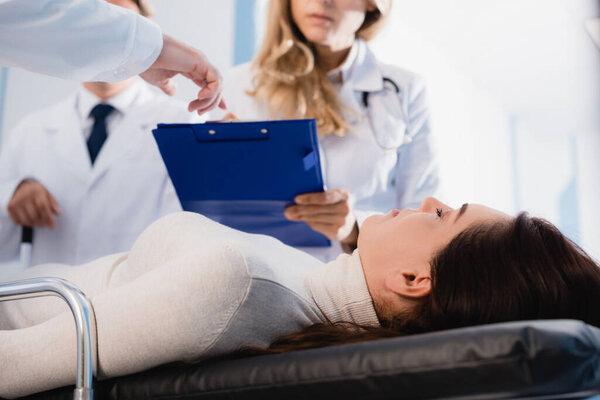 Selective focus of patient with closed eyes lying near doctor pointing at clipboard to colleague in clinic 
