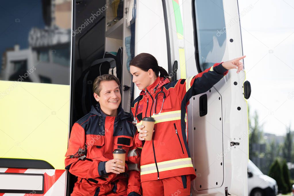 Paramedic pointing with finger while holding disposable cup near colleague and ambulance car 