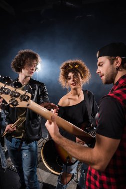 KYIV, UKRAINE - 25 AĞUSTOS 2020: Kadına yakın duran ve işaret eden kıvırcık adam, arkasında siyah ışık bulunan elektro gitarist müzisyene bakarken,