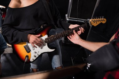 KYIV, UKRAINE - AUGUST 25, 2020: Cropped view of man touching guitar strings, while woman playing during rock band rehearsal clipart
