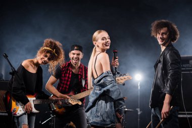 KYIV, UKRAINE - 25 AĞUSTOS 2020: Kıvırcık bateristin yanında duran mutlu rock grubu vokalisti ve arka planda arka plan ışıklandırmalı gülümseyen gitaristler