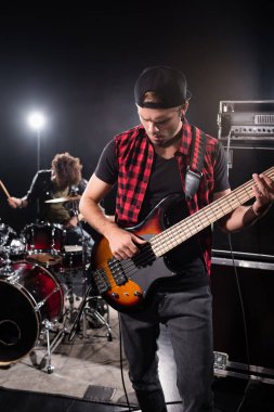 KYIV, UKRAINE - 25 AUGust 2020: Bas gitar çalan rock grubu müzisyeni, arka planda bulanık baterist ve davul setiyle birlikte karışık amplifikatörün yanında duruyor