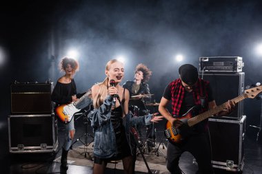 KYIV, UKRAINE - AUGUST 25, 2020: Woman with closed eyes singing while holding microphone rack near rock band musicians on blurred background clipart