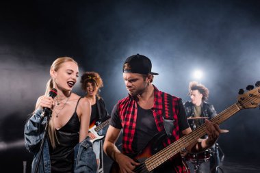KYIV, UKRAINE - 25 AĞUSTOS 2020: Rock grubunun provası sırasında arka planda bulanık bir arka plan ile gitarist yakınında şarkı söyleyen gülümseyen sarışın kadın