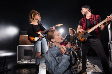 KYIV, UKRAINE - 25 Ağustos 2020: Arka planda bulanık davulcu ile gülümseyen gitaristlerin yanında otururken gözleri kapalı şarkı söyleyen kadın