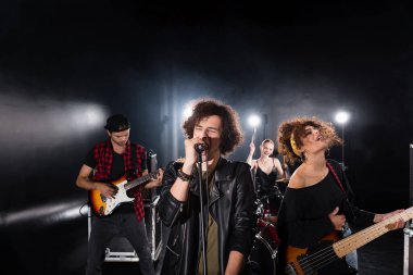 KYIV, UKRAINE - AUGUST 25, 2020: Curly vocalist with closed eyes singing in microphone near guitarists with backlit and female drummer on background clipart