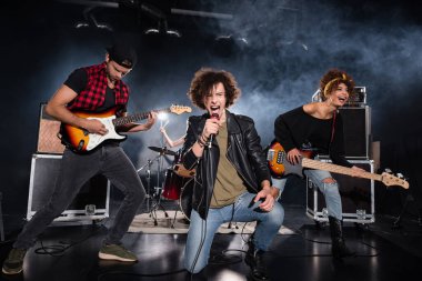 KYIV, UKRAINE - 25 AĞUSTOS 2020: Rock grubu performansı sırasında gitaristlerin yanında otururken mikrofonla bağıran kıvırcık vokalist