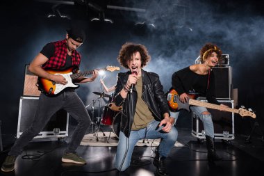 KYIV, UKRAINE - AUGUST 25, 2020: Curly vocalist shouting in microphone while sitting on knee near guitarists during rock band performance with backlit on black clipart
