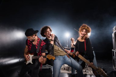 KYIV, UKRAINE - AUGUST 25, 2020: Curly vocalist singing in microphone while leaning on guitarist with backlit and blurred female drummer on background clipart